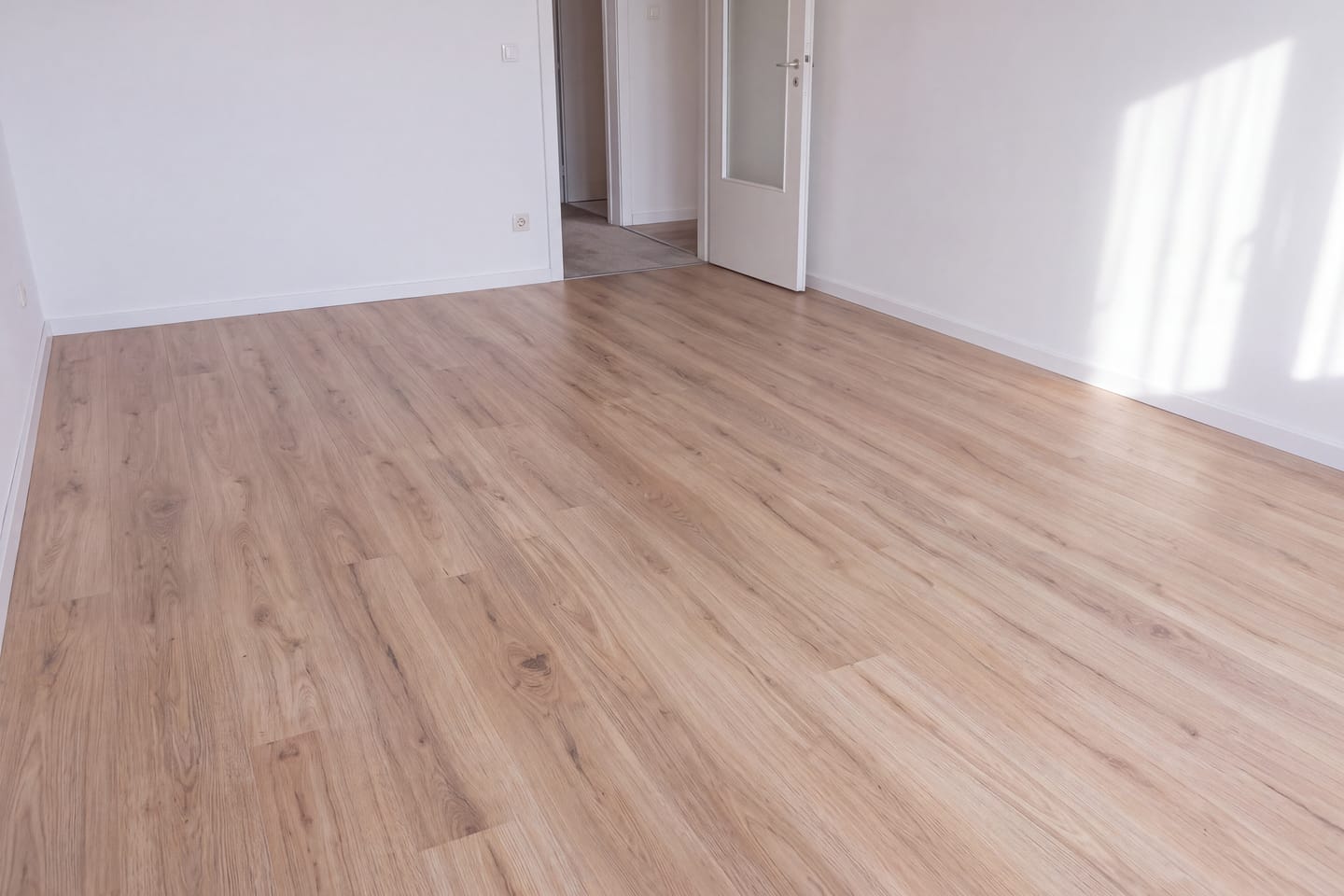 Prazna soba s novim laminatom u tonu svijetlog hrasta snimljena nisko iznad poda, bijeli zidovi, otvorena vrata prema hodniku i prijelazna lajsna na pragu